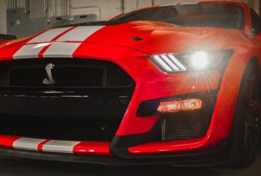 Red Ford Shelby Mustang GT500 with white racing stripes and headlights on.