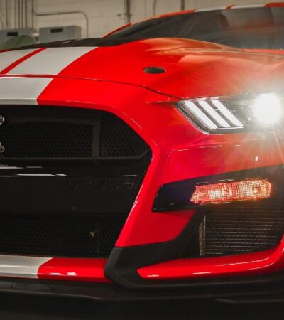 Red Ford Shelby Mustang GT500 with white racing stripes and headlights on.