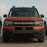 Front view of three Ford vehicles—a blue Maverick, a red-orange Bronco Sport, and a white Mustang Mach-E—parked side by side on a wide open asphalt surface under a partly cloudy sky.