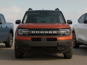 Front view of three Ford vehicles—a blue Maverick, a red-orange Bronco Sport, and a white Mustang Mach-E—parked side by side on a wide open asphalt surface under a partly cloudy sky.