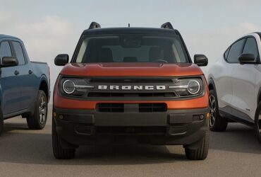 Front view of three Ford vehicles—a blue Maverick, a red-orange Bronco Sport, and a white Mustang Mach-E—parked side by side on a wide open asphalt surface under a partly cloudy sky.
